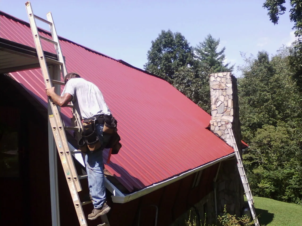 Roofer on a ladder beside a red metal roof and stone chimney — Soffit & Fascia Repair by Roofing Brothers in Okmulgee